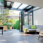 A bright and airy sunroom interior featuring a large sectional sofa and glass walls that fold open to a landscaped patio.