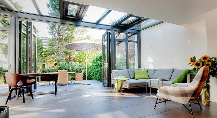 A bright and airy sunroom interior featuring a large sectional sofa and glass walls that fold open to a landscaped patio.
