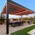 A freestanding motorized pergola with red LED mood lighting installed over a modern outdoor kitchen, pizza oven, and large dining table.