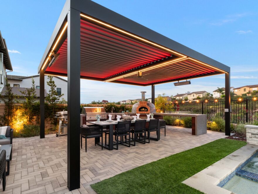 A freestanding motorized pergola with red LED mood lighting installed over a modern outdoor kitchen, pizza oven, and large dining table.