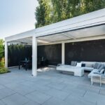 A white freestanding pergola with retractable privacy screens covers a stone paver patio, creating a dedicated dining area and a separate lounge zone with a sectional sofa and marble-style coffee table.