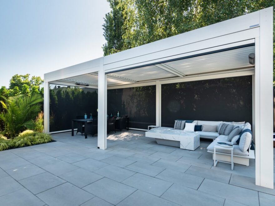 A white freestanding pergola with retractable privacy screens covers a stone paver patio, creating a dedicated dining area and a separate lounge zone with a sectional sofa and marble-style coffee table.