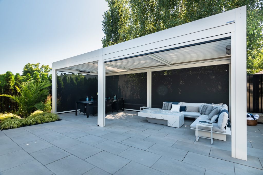 A white freestanding pergola with retractable privacy screens covers a stone paver patio, creating a dedicated dining area and a separate lounge zone with a sectional sofa and marble-style coffee table.