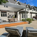 A light gray aluminum louvered pergola in the open position casts striped sunlight over a wood deck and teak dining set, attached to a large stucco estate home with a brick paver patio and pool just steps away.