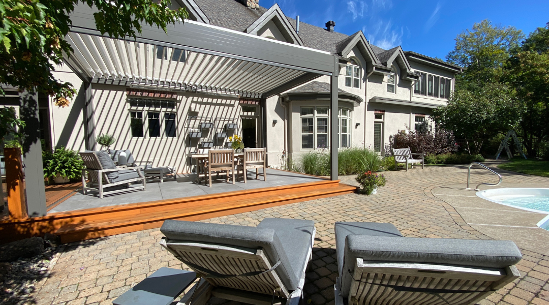 A light gray aluminum louvered pergola in the open position casts striped sunlight over a wood deck and teak dining set, attached to a large stucco estate home with a brick paver patio and pool just steps away.